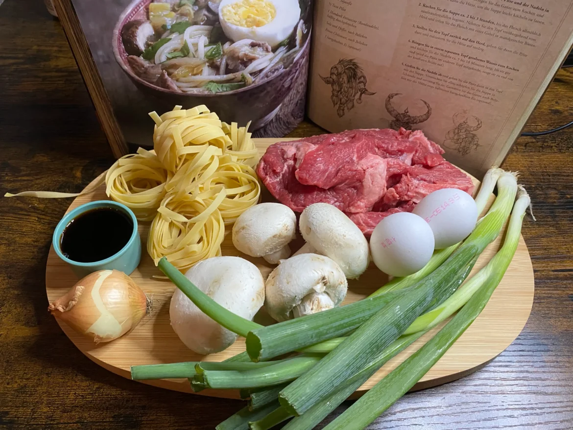 Yakbrühe-Zutaten: Rindfleisch, Fettucine, hartgekochte Eier, Pilze, Lauch, Sojasoße, Zwiebel und Knoblauch auf einem Holztisch arrangiert.